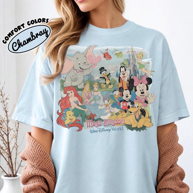 May include: Light blue t-shirt featuring a graphic of Disney characters, including Dumbo, Ariel, Mickey Mouse, and Minnie Mouse. The shirt has the words "Magic Kingdom Walt Disney World" printed on it. The shirt also has the words "Comfort Colors Chambray" printed on it.