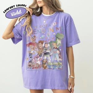 May include: A violet short-sleeve t-shirt featuring a graphic print of various animated toy characters and the text "YOU'VE GOT A FRIEND IN ME". The design includes characters like a cowboy, space ranger, cowgirl, dinosaur, and aliens. A small logo reads "COMFORT COLORS Violet".