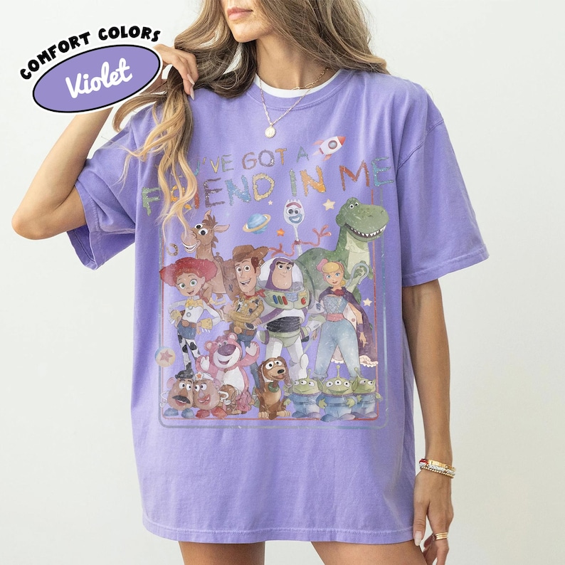 May include: A violet short-sleeve t-shirt featuring a graphic print of various animated toy characters and the text "YOU'VE GOT A FRIEND IN ME". The design includes characters like a cowboy, space ranger, cowgirl, dinosaur, and aliens. A small logo reads "COMFORT COLORS Violet".