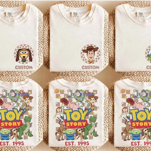 Puede incluir: Camisetas blanquecinas con personajes de Toy Story. Algunas camisetas tienen retratos de personajes con el texto "CUSTOM", mientras que otras muestran un gr&aacute;fico colorido con el texto "TOY STORY EST. 1995". Las camisetas est&aacute;n dobladas y colocadas sobre una superficie tejida.