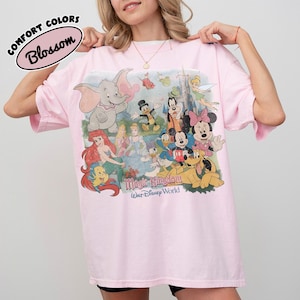 May include: A light pink t-shirt with a graphic featuring Disney characters like Dumbo, Mickey Mouse, and Ariel. The shirt has the words "Magic Kingdom" and "Walt Disney World" printed on it. The text "Comfort Colors Blossom" is also printed on the shirt.