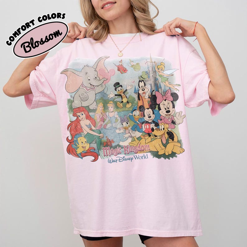 May include: A light pink t-shirt with a graphic featuring Disney characters like Dumbo, Mickey Mouse, and Ariel. The shirt has the words "Magic Kingdom" and "Walt Disney World" printed on it. The text "Comfort Colors Blossom" is also printed on the shirt.