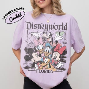 May include: A light purple t-shirt with a Disney World graphic featuring Mickey Mouse, Minnie Mouse, Donald Duck, Daisy Duck, Goofy, and Pluto. The shirt has the text "Disneyworld Florida" and "EST. 1971". The shirt is labeled "Comfort Colors Orchid".