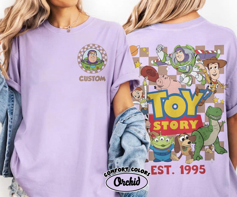 Puede incluir: Una camiseta de color lila claro con Buzz Lightyear de Toy Story. La parte delantera de la camiseta tiene un gr&aacute;fico circular con Buzz Lightyear y la palabra "CUSTOM". La parte trasera de la camiseta tiene un gr&aacute;fico m&aacute;s grande con varios personajes de Toy Story y las palabras "TOY STORY EST. 1995".