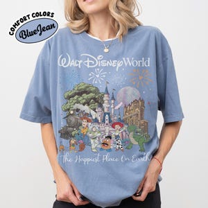 May include: Blue short-sleeved t-shirt with a graphic of Walt Disney World, featuring a castle, characters, and fireworks. The text "The Happiest Place On Earth" is below the graphic. The shirt has the words "Comfort Colors" and "Blue Jean" in a circle.