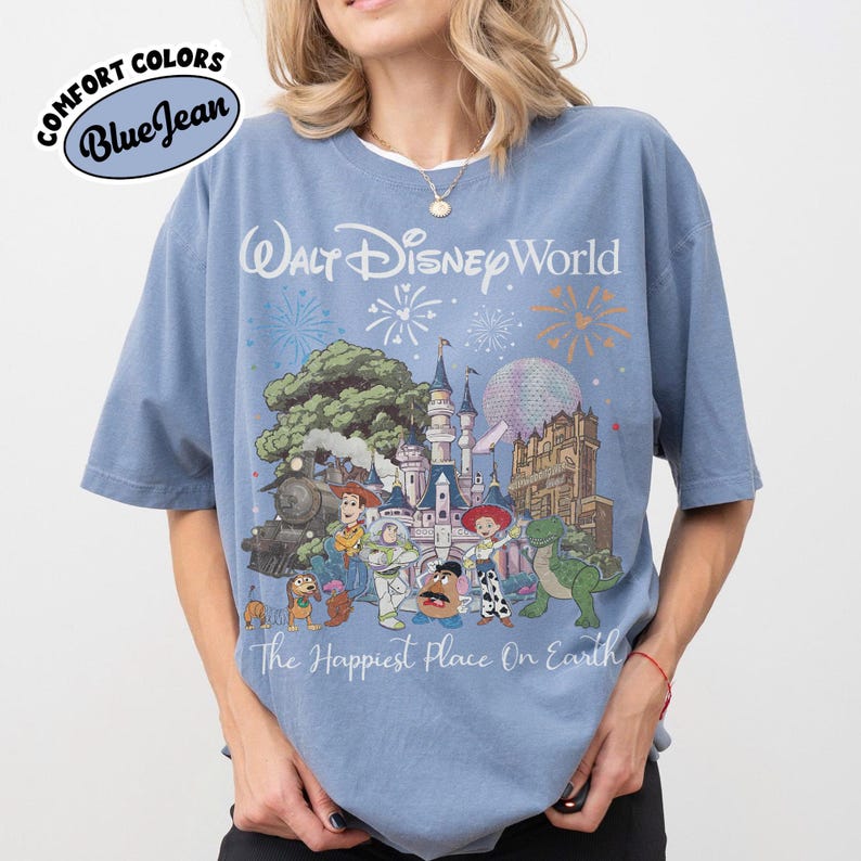 May include: Blue short-sleeved t-shirt with a graphic of Walt Disney World, featuring a castle, characters, and fireworks. The text "The Happiest Place On Earth" is below the graphic. The shirt has the words "Comfort Colors" and "Blue Jean" in a circle.