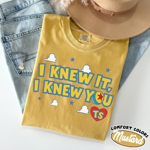 May include: A mustard yellow short-sleeve t-shirt featuring the blue text "I KNEW IT, I KNEW YOU" with a red heart containing the letters "TS" and a small ball graphic. The shirt includes white cloud and blue star accents, with a Comfort Colors label.