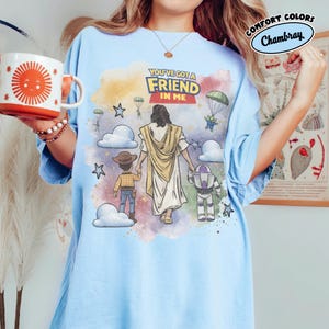 May include: Light blue t-shirt with a graphic featuring Jesus walking with cartoon characters, including Woody and Buzz Lightyear. The text "YOU'VE GOT A FRIEND IN ME" is displayed above the figures. The shirt has the words "COMFORT COLORS" and "Chambray" on the upper right.