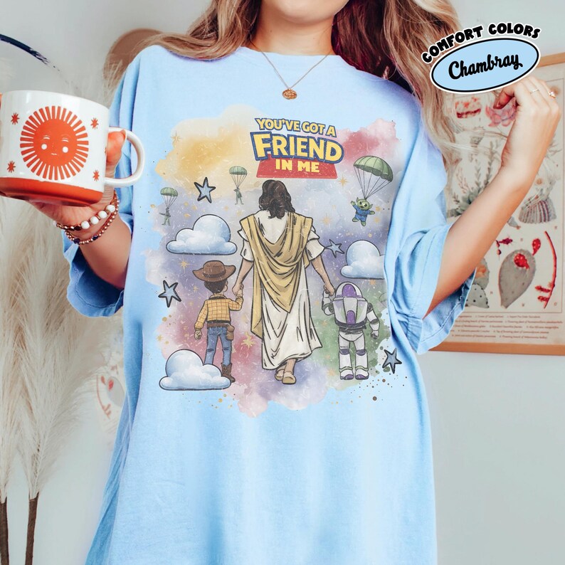 May include: Light blue t-shirt with a graphic featuring Jesus walking with cartoon characters, including Woody and Buzz Lightyear. The text "YOU'VE GOT A FRIEND IN ME" is displayed above the figures. The shirt has the words "COMFORT COLORS" and "Chambray" on the upper right.