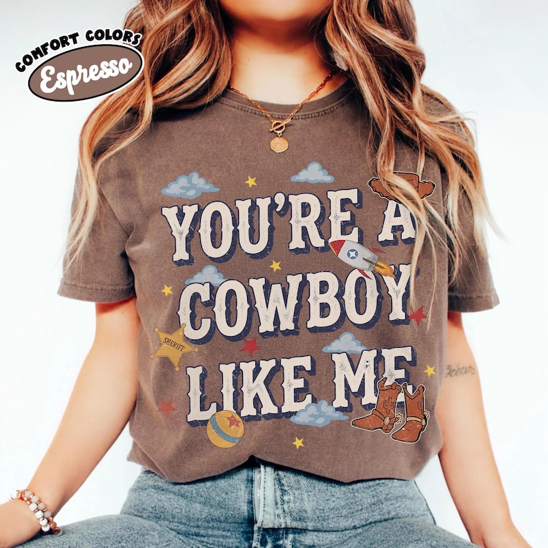 May include: An espresso-colored short-sleeve t-shirt featuring the text "YOU'RE A COWBOY LIKE ME" in a large, light-colored font. The design includes graphic illustrations of a rocket, a sheriff star, a ball, and cowboy boots with clouds and stars.