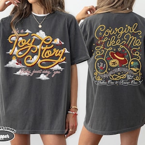 以下が含まれることがあります： チャコールグレーのTシャツ。フロントには白い雲の中に黄色いロープ風の文字で「it's a Toy Story」とプリントされています。バックには赤い帽子、ロープ、音符と共に「Cowgirl like me」という文字がデザインされています。