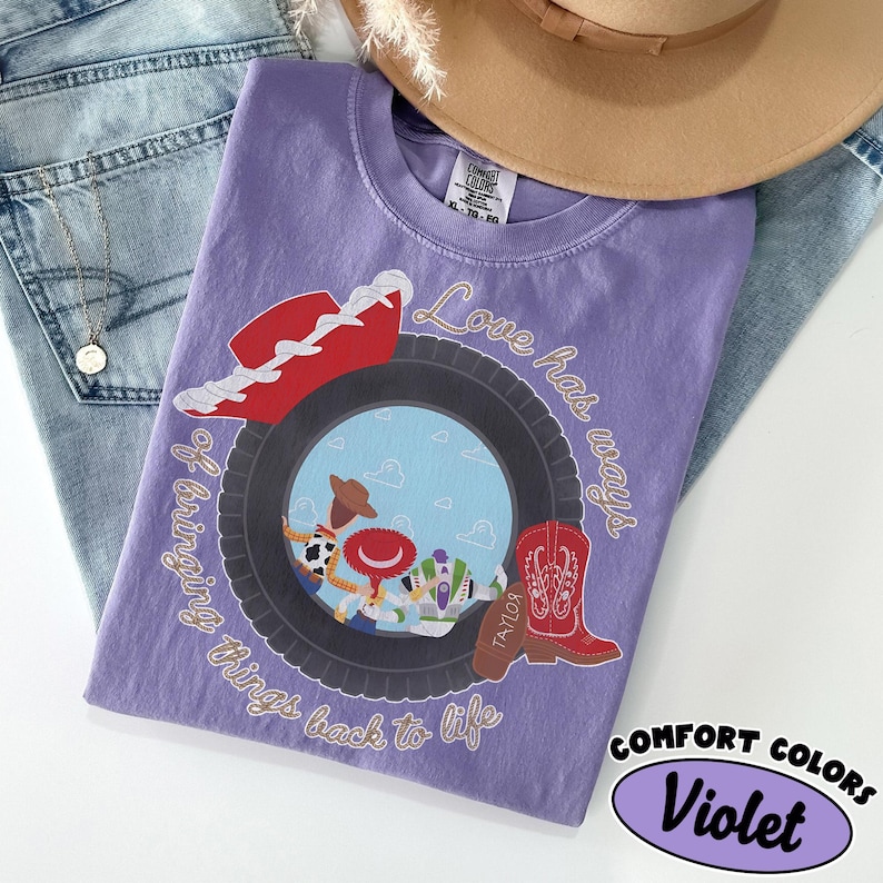 May include: A violet short-sleeved t-shirt featuring a graphic of a black tire framing cartoon characters, a red cowboy hat, and a red boot. The text reads Love has ways of bringing things back to life. The shirt has a Comfort Colors brand label.
