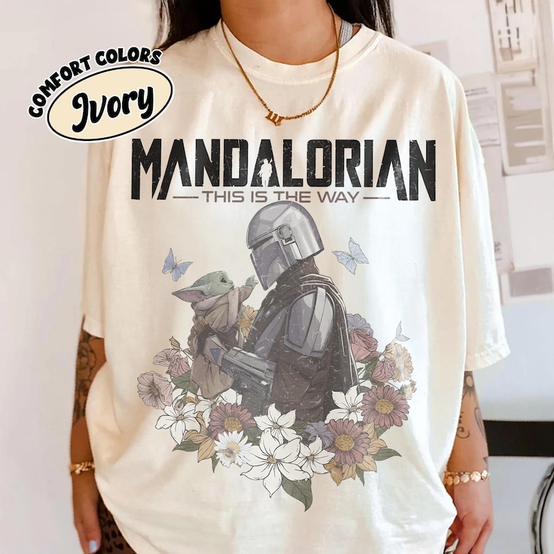 May include: Ivory-colored t-shirt with a graphic featuring The Mandalorian and Grogu surrounded by flowers and butterflies. The text "MANDALORIAN - THIS IS THE WAY" is printed above the image. The shirt has a Comfort Colors label.