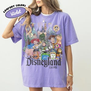 May include: A violet short-sleeve t-shirt featuring a graphic print of various Toy Story characters in front of a Disneyland castle. The text reads COMFORT COLORS Violet, Disneyland, and Est. 1955. The design includes Woody, Buzz Lightyear, Jessie, Rex, Hamm, Mr. Potato Head, and Slinky Dog.