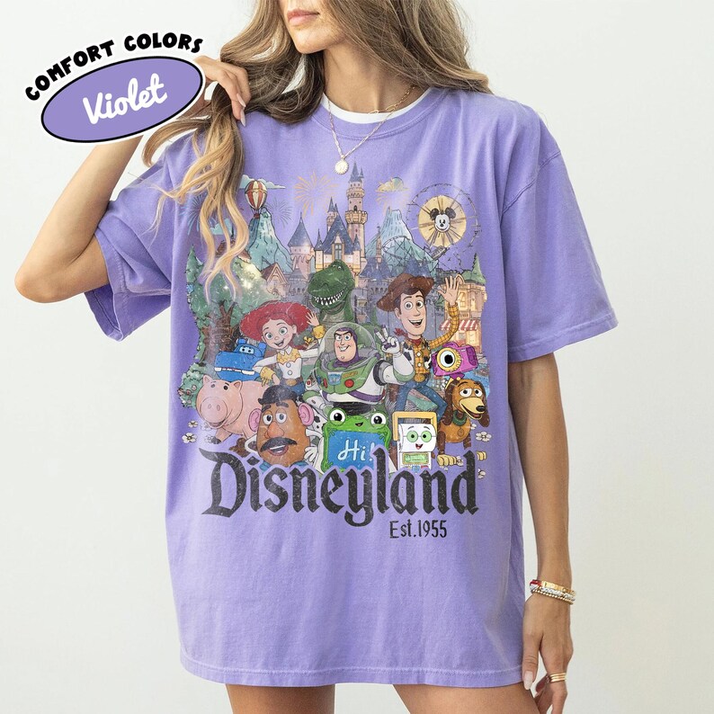 May include: A violet short-sleeve t-shirt featuring a graphic print of various Toy Story characters in front of a Disneyland castle. The text reads COMFORT COLORS Violet, Disneyland, and Est. 1955. The design includes Woody, Buzz Lightyear, Jessie, Rex, Hamm, Mr. Potato Head, and Slinky Dog.