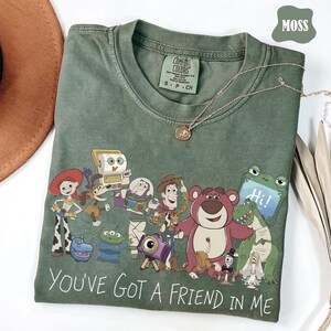 May include: A moss green short-sleeved t-shirt featuring a graphic print of various animated characters from the Toy Story franchise. The text "YOU'VE GOT A FRIEND IN ME" is printed in white at the bottom. The shirt has a crew neckline and a tag.