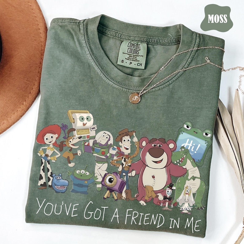 May include: A moss green short-sleeved t-shirt featuring a graphic print of various animated characters from the Toy Story franchise. The text "YOU'VE GOT A FRIEND IN ME" is printed in white at the bottom. The shirt has a crew neckline and a tag.