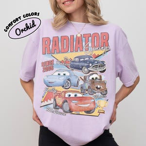 May include: A light purple t-shirt featuring a graphic of characters from the movie Cars, including Lightning McQueen, Sally, Mater, and Doc Hudson. The shirt has the words "Radiator Springs" and "Since 2006" printed on it.