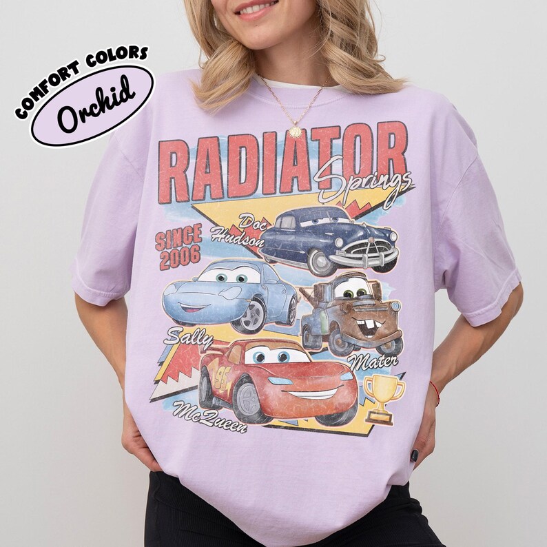 May include: A light purple t-shirt featuring a graphic of characters from the movie Cars, including Lightning McQueen, Sally, Mater, and Doc Hudson. The shirt has the words "Radiator Springs" and "Since 2006" printed on it.