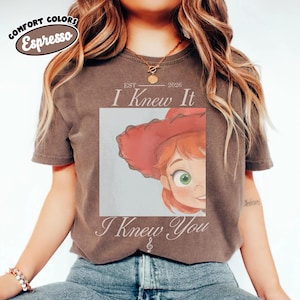 Op de afbeelding: Een donkerbruin t-shirt met korte mouwen en een afbeelding van een stripfiguur met rood haar en een rode hoed. De tekst luidt "EST 2026 I Knew It I Knew You" met een kleine muzieknoot. Het shirt heeft een Comfort Colors Espresso label.