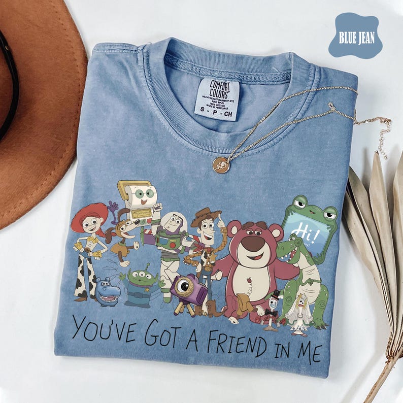 May include: A blue jean colored t-shirt featuring a graphic print of various animated characters from the Toy Story franchise. The text "YOU'VE GOT A FRIEND IN ME" is printed in black below the characters. The shirt has a crew neckline and a tag.
