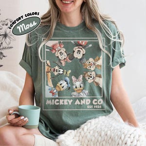 Puede incluir: Camiseta verde musgo con un gr&aacute;fico de Mickey Mouse, Minnie Mouse, Pato Donald, Daisy, Goofy y Chip y Chop. El texto "Mickey and Co. Est. 1928" est&aacute; impreso debajo. La camiseta tiene mangas enrolladas.