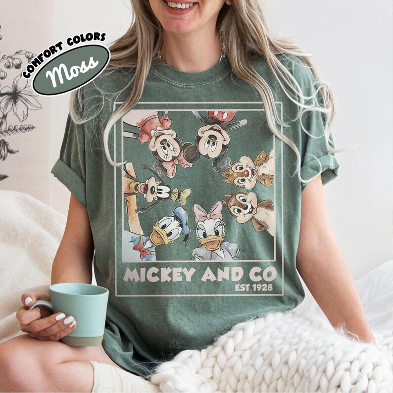 Puede incluir: Camiseta verde musgo con un gr&aacute;fico de Mickey Mouse, Minnie Mouse, Pato Donald, Daisy, Goofy y Chip y Chop. El texto "Mickey and Co. Est. 1928" est&aacute; impreso debajo. La camiseta tiene mangas enrolladas.