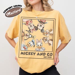 Puede incluir: Camiseta amarilla mostaza con un gr&aacute;fico enmarcado de Mickey Mouse, Minnie Mouse, Donald Duck, Daisy Duck, Goofy, Chip y Dale. El texto "Mickey and Co. Est. 1928" est&aacute; impreso debajo. La camiseta tiene mangas cortas y cuello redondo.