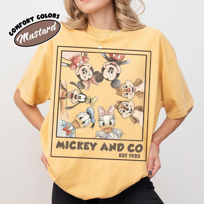 Puede incluir: Camiseta amarilla mostaza con un gr&aacute;fico enmarcado de Mickey Mouse, Minnie Mouse, Donald Duck, Daisy Duck, Goofy, Chip y Dale. El texto "Mickey and Co. Est. 1928" est&aacute; impreso debajo. La camiseta tiene mangas cortas y cuello redondo.