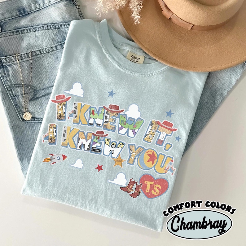 May include: A light blue t-shirt featuring the text I KNEW IT, I KNEW YOU in a multi-colored font with patterns inspired by cowboy and space characters. The design includes small stars, clouds, a rocket, and a red heart with the letters TS inside.
