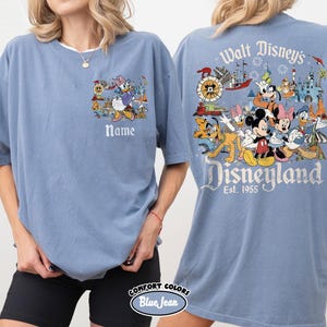 Peut inclure: Un t-shirt bleu Comfort Colors avec un motif Disneyland. Le devant pr&eacute;sente Daisy Duck et le mot "Name". Le dos affiche Mickey Mouse, Minnie Mouse et d'autres personnages Disney avec les mots "Walt Disney's Disneyland Est. 1955".