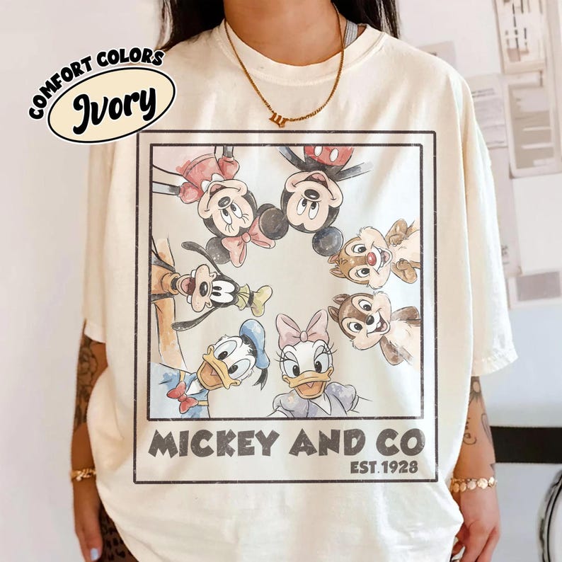 Puede incluir: Camiseta de color marfil con un gr&aacute;fico circular de personajes de Disney, incluyendo Mickey y Minnie Mouse, Donald y Daisy Duck, Goofy y Chip y Chop. El gr&aacute;fico est&aacute; enmarcado con el texto "Mickey and Co. EST.1928". La camiseta tiene "Comfort Colors" impreso.