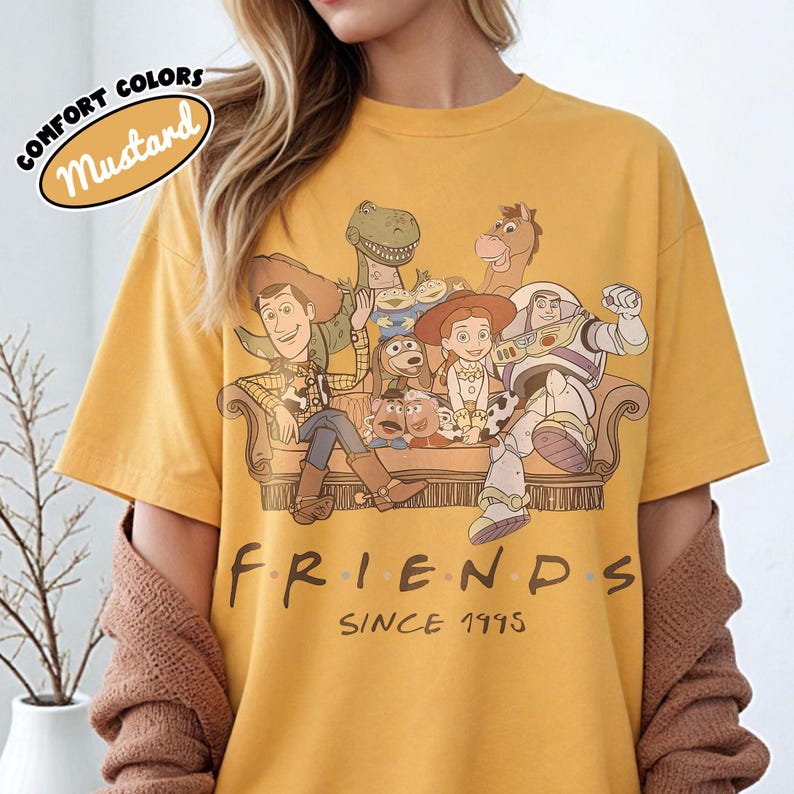 May include: Mustard yellow t-shirt featuring a graphic of Toy Story characters on a couch. The word "FRIENDS" is printed below the graphic, with "SINCE 1995" underneath. The shirt has short sleeves and a crew neck.