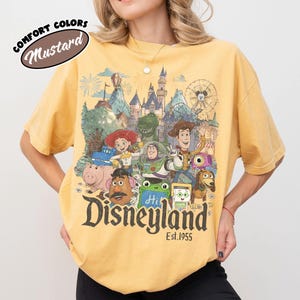 May include: A mustard yellow t-shirt featuring a graphic print of various animated characters in front of a castle and a Ferris wheel. The text reads "Disneyland Est. 1955" in a dark font at the bottom. A small oval label in the corner says "Comfort Colors Mustard".