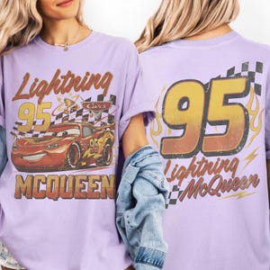 Może przedstawiać: Jasnofioletowa koszulka z grafiką Zygzaka McQueena z filmu Auta. Prz&oacute;d koszulki przedstawia napis "Lightning 95 McQueen" z wizerunkiem samochodu. Tył ma duży napis "95" z płomieniami i "Lightning McQueen".