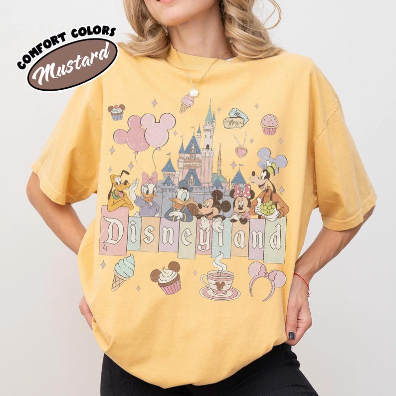 May include: A mustard yellow t-shirt featuring a graphic print of a castle and various cartoon characters including Mickey Mouse, Minnie Mouse, Donald Duck, Daisy Duck, Goofy, and Pluto. The text Disneyland is printed across the center.