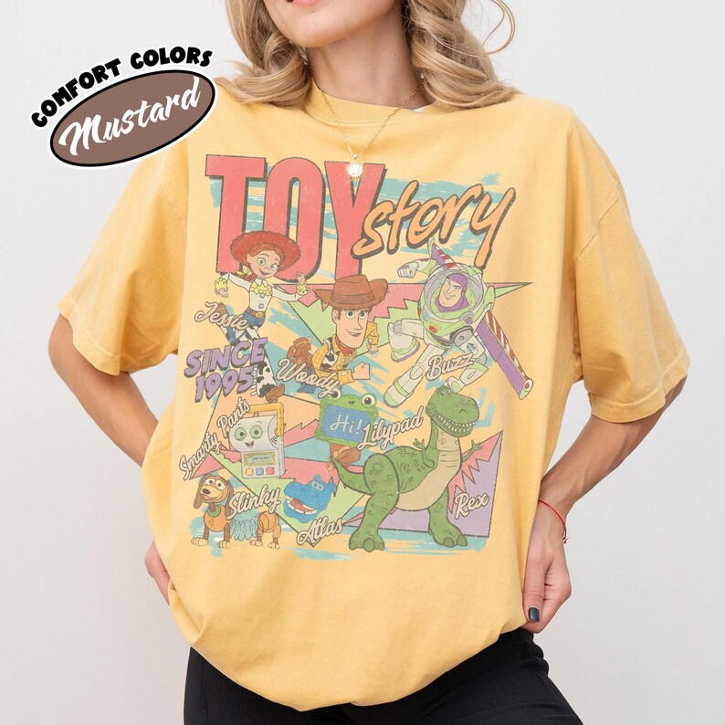 May include: A mustard yellow t-shirt featuring a graphic print of Toy Story characters including Woody, Buzz Lightyear, Jessie, Rex, Slinky Dog, and others. The design includes the text "TOY story", "SINCE 1995", and character names in a colorful, retro style.