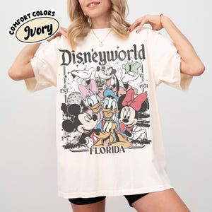 May include: Ivory-colored t-shirt featuring a graphic of Disney characters, including Mickey and Minnie Mouse, Donald and Daisy Duck, and Goofy. The shirt has the text "Disneyworld Florida" and "EST. 1971". The shirt has the words "Comfort Colors" and "Ivory" in a circle.