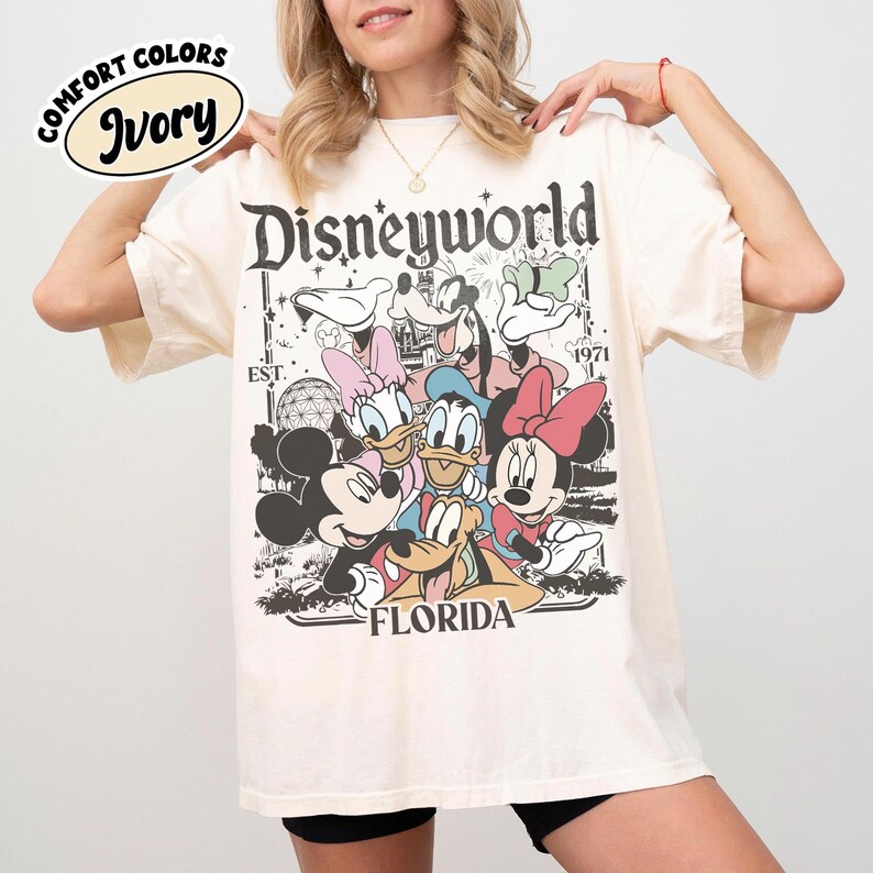 May include: Ivory-colored t-shirt featuring a graphic of Disney characters, including Mickey and Minnie Mouse, Donald and Daisy Duck, and Goofy. The shirt has the text "Disneyworld Florida" and "EST. 1971". The shirt has the words "Comfort Colors" and "Ivory" in a circle.