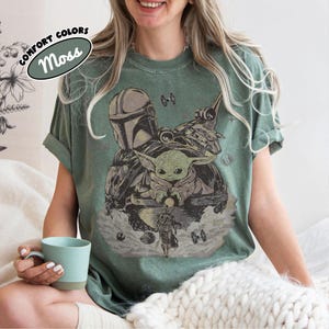 Puede incluir: Camiseta verde musgo con un gr&aacute;fico de The Mandalorian, Grogu y una nave espacial. El dise&ntilde;o incluye el texto "COMFORT COLORS" y "Moss" en un &oacute;valo. La camiseta tiene un corte relajado.