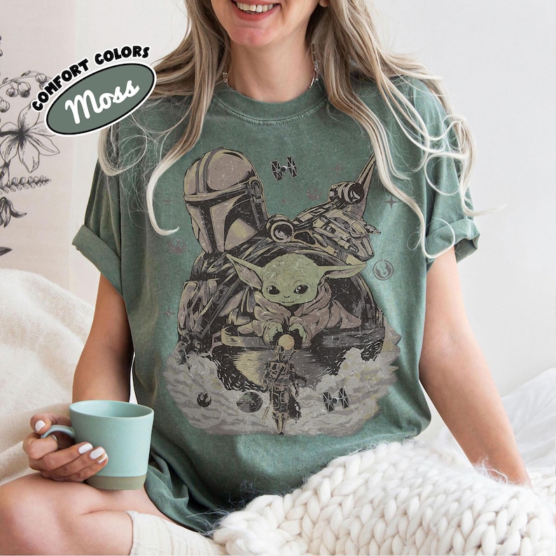 Puede incluir: Camiseta verde musgo con un gr&aacute;fico de The Mandalorian, Grogu y una nave espacial. El dise&ntilde;o incluye el texto "COMFORT COLORS" y "Moss" en un &oacute;valo. La camiseta tiene un corte relajado.
