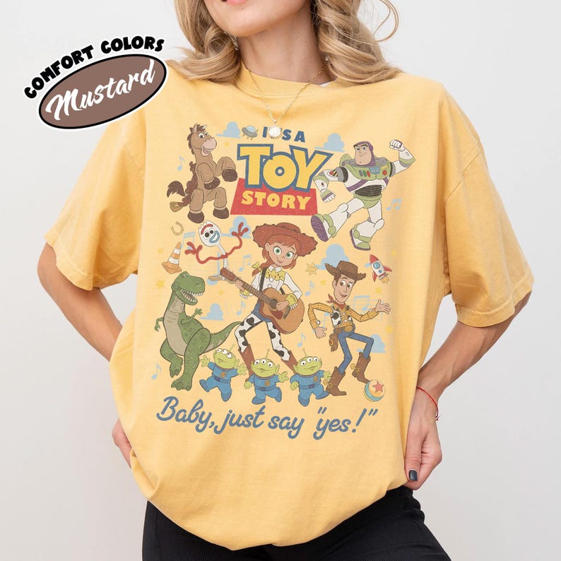 May include: A mustard yellow short-sleeve t-shirt featuring a graphic print of Toy Story characters including Woody, Jessie, Buzz Lightyear, Rex, Bullseye, Forky, and the aliens. The text reads "TOY STORY" and "Baby, just say 'yes!'".