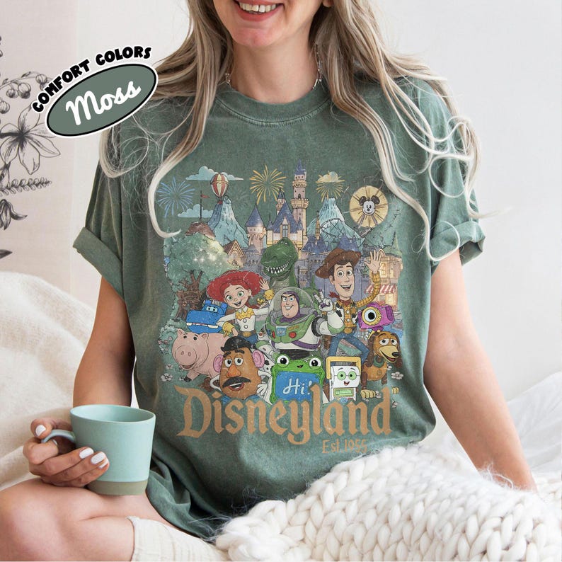 May include: A moss green short-sleeve t-shirt featuring a graphic print of various animated characters in front of a castle and fireworks. The text "Disneyland Est. 1955" is printed in gold at the bottom. A small label reads "Comfort Colors Moss".