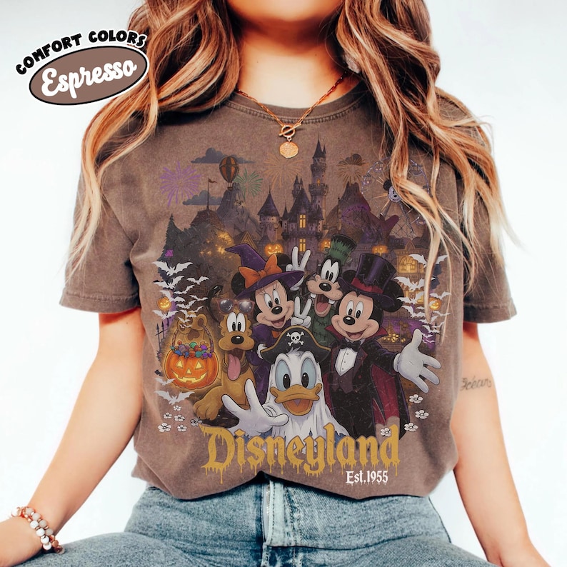 May include: An espresso-colored t-shirt featuring a graphic of Disney characters in Halloween costumes, including Mickey Mouse as a vampire, Minnie Mouse as a witch, and Donald Duck as a ghost, with the text Disneyland Est. 1955 and Comfort Colors Espresso.