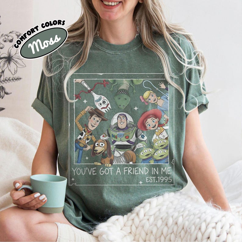 May include: Moss green t-shirt featuring a graphic of characters from a popular animated film. The design includes Woody, Buzz Lightyear, and other characters, with the text "You've Got a Friend in Me" and "EST.1995". The shirt has rolled-up sleeves.