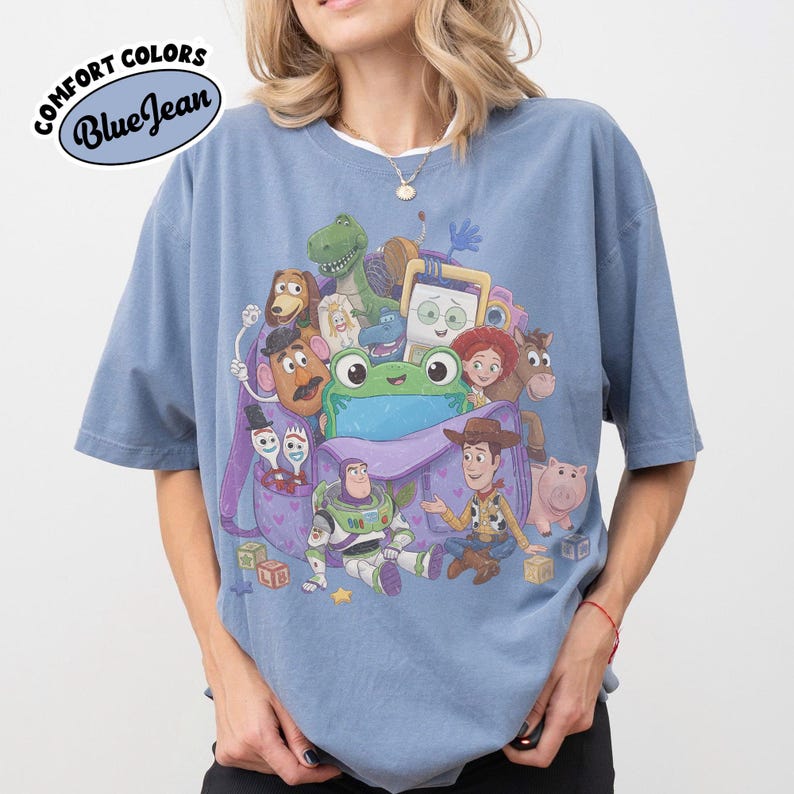 Pode incluir: Uma t-shirt azul de manga curta com uma impress&atilde;o gr&aacute;fica de v&aacute;rias personagens animadas de Toy Story, incluindo Woody, Buzz Lightyear e um sapo verde central, a sair de uma mochila roxa. O texto Comfort Colors Blue Jean &eacute; vis&iacute;vel.