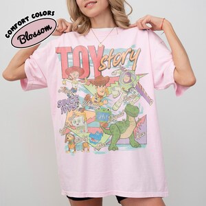 May include: A light pink t-shirt featuring a graphic print of Toy Story characters including Woody, Buzz Lightyear, Jessie, Rex, Slinky, and others. The text reads Toy Story, Since 1995, Jessie, Woody, Buzz, Smarty Pants, Lilypad, Slinky, and Rex.