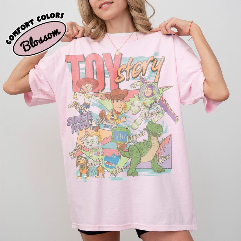 May include: A light pink t-shirt featuring a graphic print of Toy Story characters including Woody, Buzz Lightyear, Jessie, Rex, Slinky, and others. The text reads Toy Story, Since 1995, Jessie, Woody, Buzz, Smarty Pants, Lilypad, Slinky, and Rex.