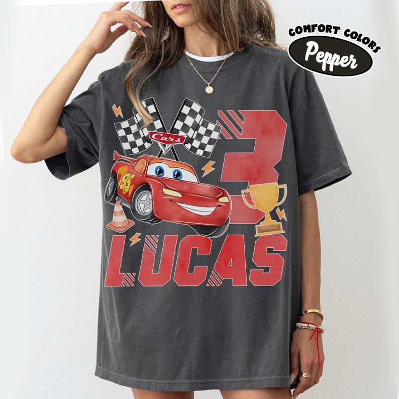 Puede incluir: Camiseta gris oscuro con un coche de carreras rojo de dibujos animados, el n&uacute;mero 95 y el nombre "LUCAS" en rojo. Tambi&eacute;n se muestran banderas a cuadros, el n&uacute;mero 3 y un trofeo dorado. La camiseta tiene las palabras "Comfort Colors Pepper" impresas.