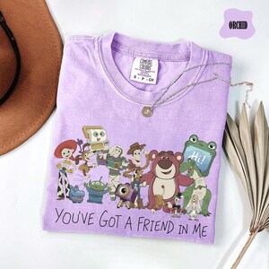 May include: An orchid-colored t-shirt featuring a graphic print of various animated characters from the Toy Story franchise. Below the characters, the text reads, "YOU'VE GOT A FRIEND IN ME." The shirt has a crew neckline and a Comfort Colors brand label.